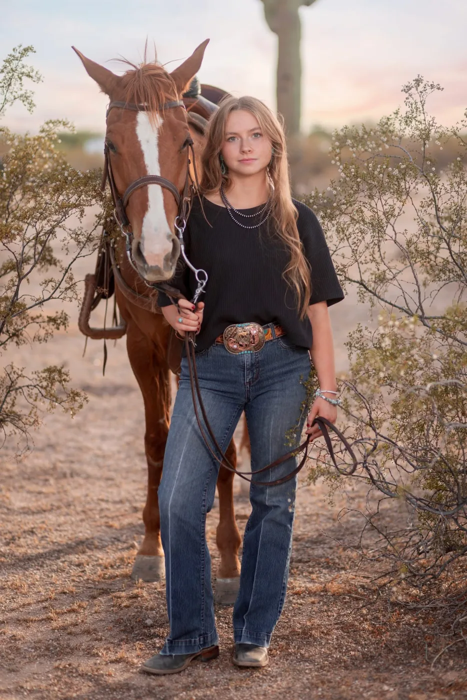 Senior portrait of a young woman leading her chestnut horse through the desert at sunset, by Casa de Baca Studios