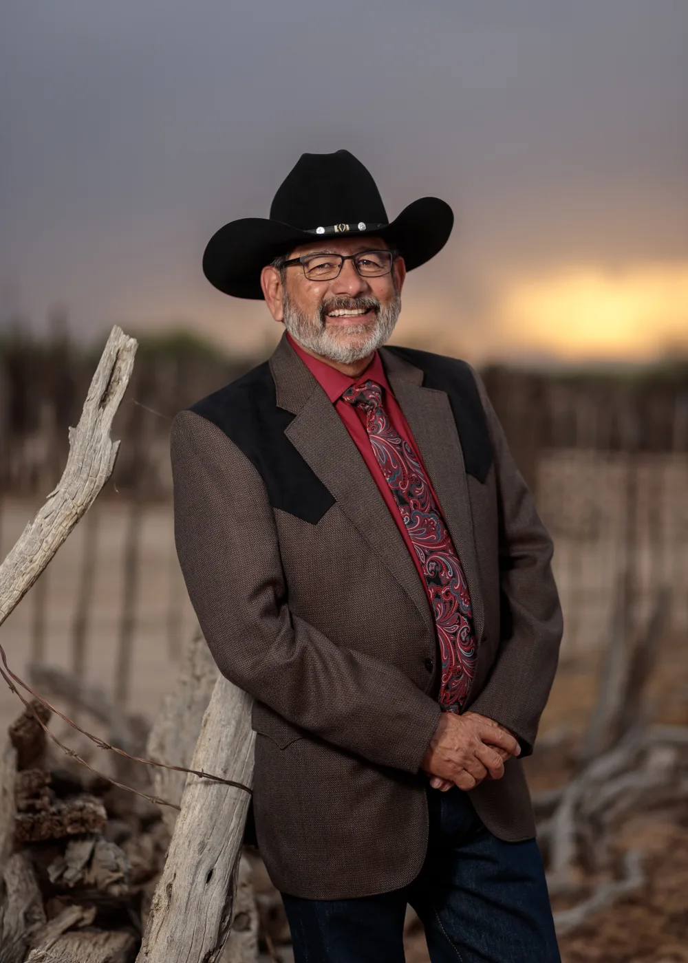 Portrait of a man in a black cowboy hat and western jacket leaning on a weathered fence post at sunset, by Casa de Baca Studios